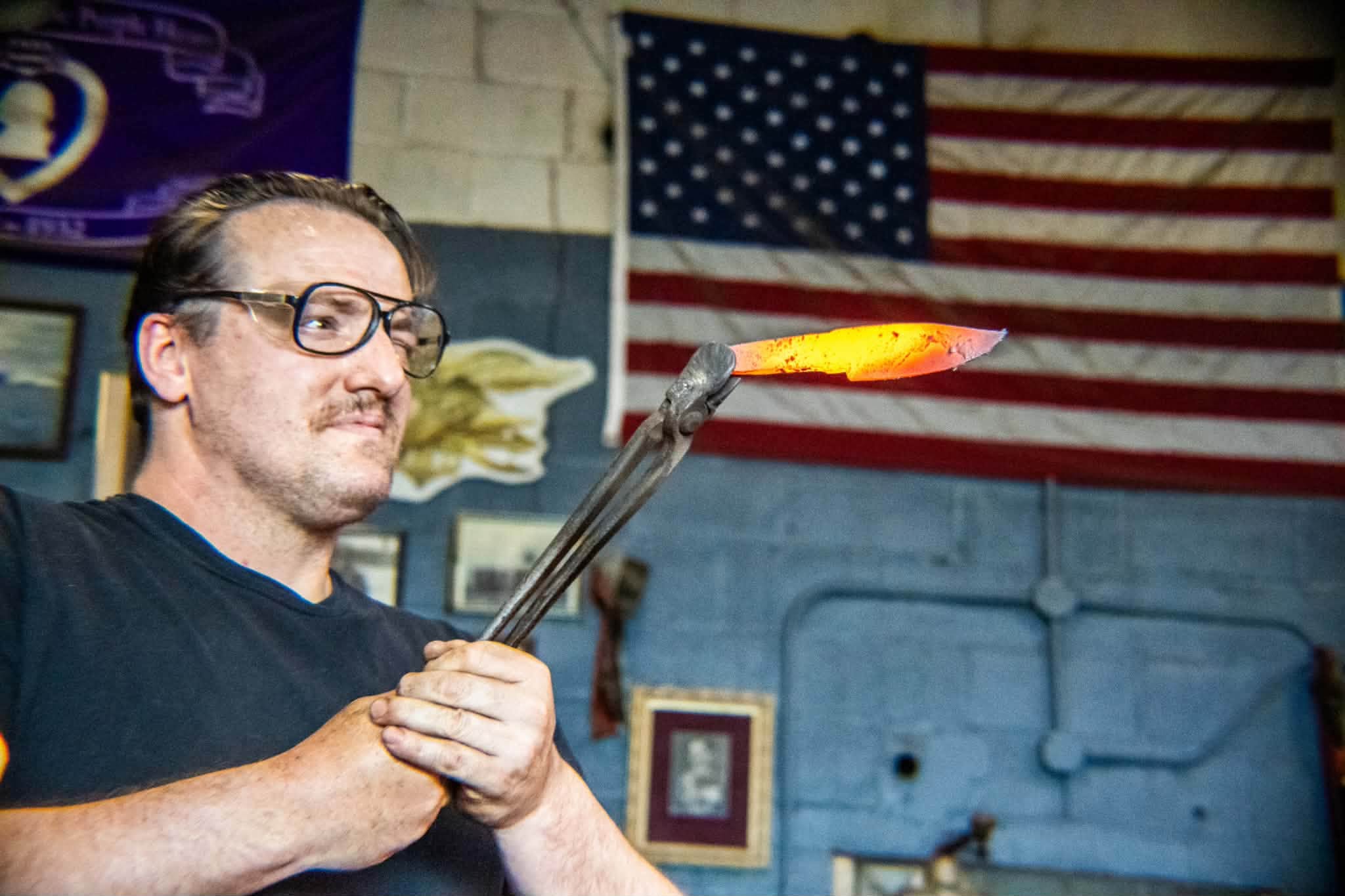 Seth Boehnke holding a glowing blade in tongs, an American flag on the wall behind him