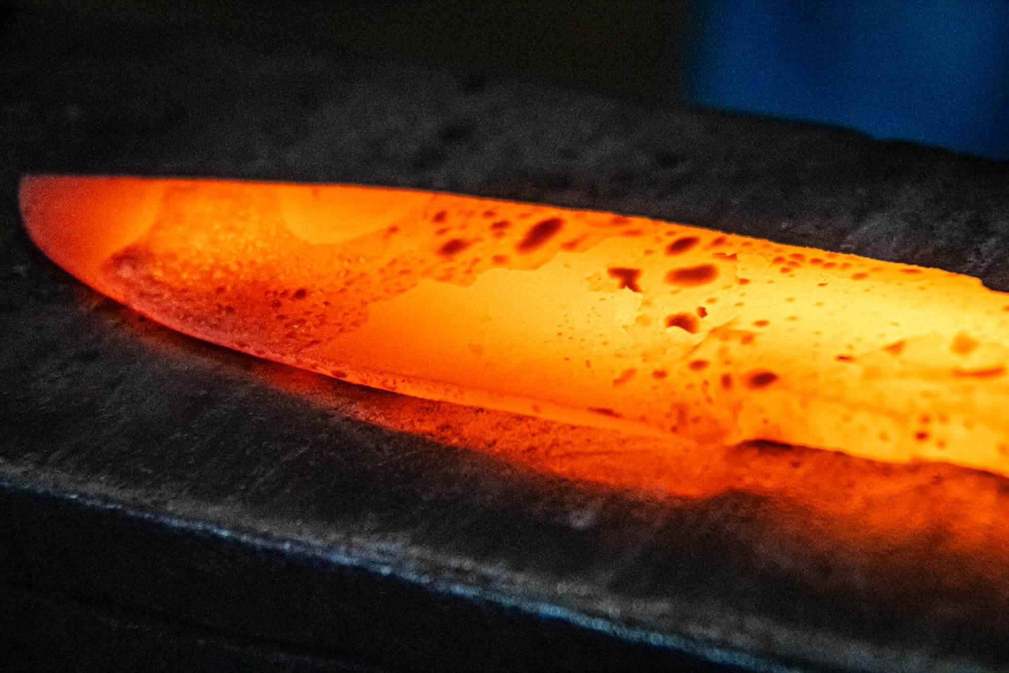 Close-up of a blade glowing orange on the anvil, scale flaking from the surface