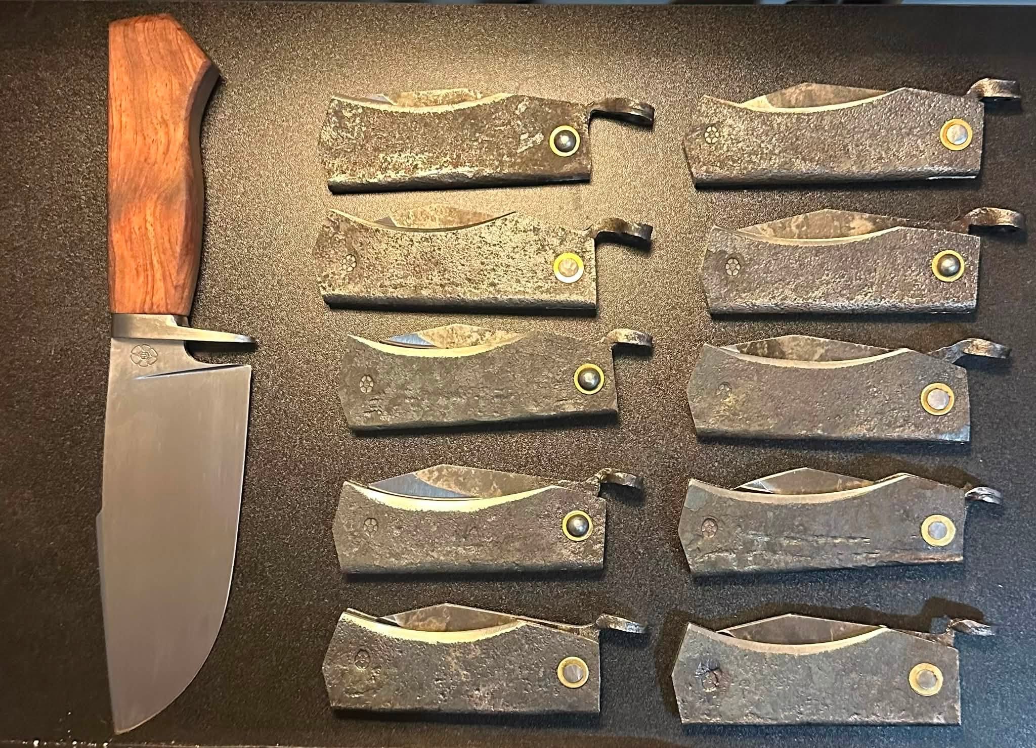 Ten forged slipjoint folders laid out in two rows beside a fixed blade with a hardwood handle