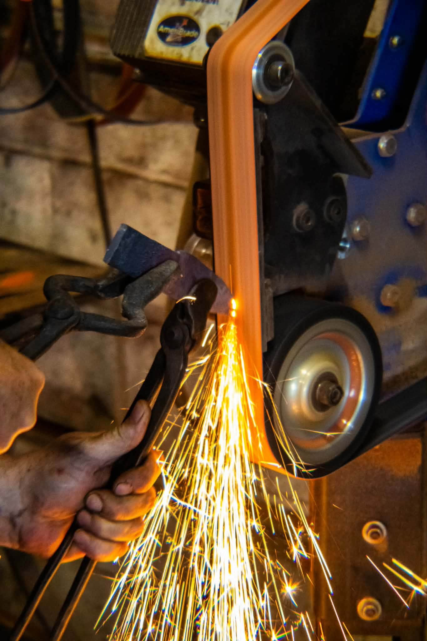 A blade held in tongs against a belt grinder, sparks streaming off the edge